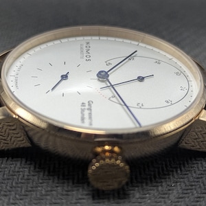 Pu&ograve; includere: Un orologio da polso color oro con quadrante bianco, lancette blu sottili e cinturino in pelle marrone. Il quadrante riporta la scritta NOMOS GLASHUTTE, MADE IN GERMANY e Gangreserve 48 Stunden. La cassa presenta un motivo testurizzato.
