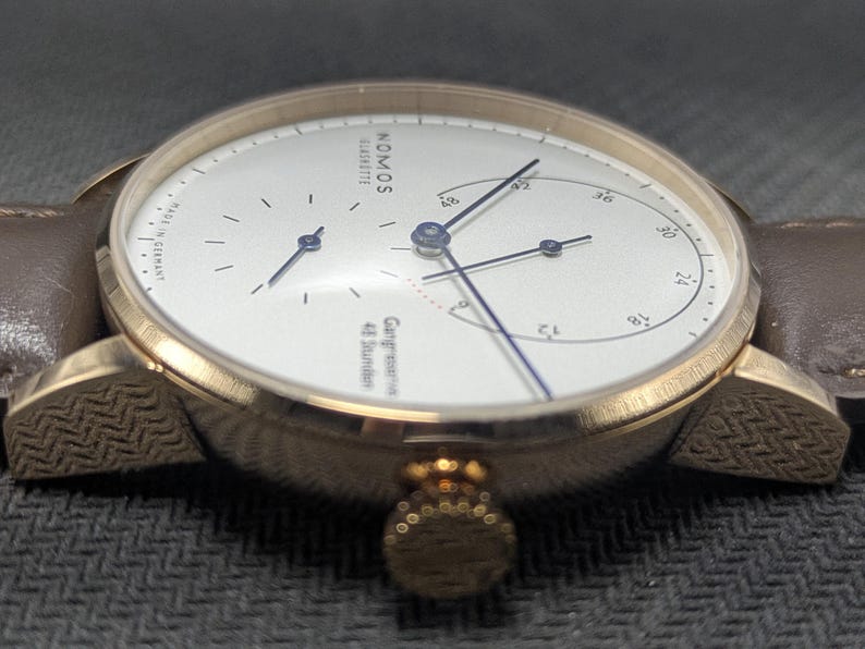 Pu&ograve; includere: Un orologio da polso color oro con quadrante bianco, lancette blu sottili e cinturino in pelle marrone. Il quadrante riporta la scritta NOMOS GLASHUTTE, MADE IN GERMANY e Gangreserve 48 Stunden. La cassa presenta un motivo testurizzato.