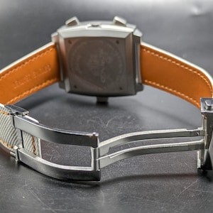 May include: A square-cased wristwatch featuring a silver-tone metal case with two pushers and a crown. The strap is light grey with a tan leather lining and includes a silver-tone metal deployment clasp mechanism visible in the open position.