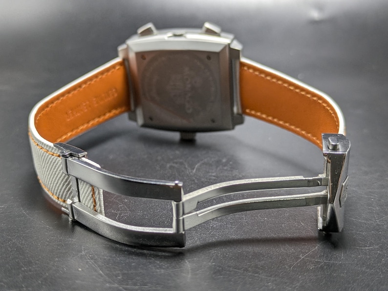 May include: A square-cased wristwatch featuring a silver-tone metal case with two pushers and a crown. The strap is light grey with a tan leather lining and includes a silver-tone metal deployment clasp mechanism visible in the open position.
