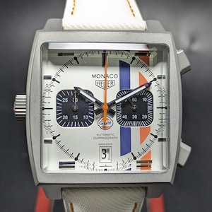 May include: A square-cased TAG Heuer Monaco chronograph watch with a silver dial, featuring two black sub-dials, a date window at the six o'clock position, and vertical blue and orange racing stripes. The watch has a white strap and orange hands.