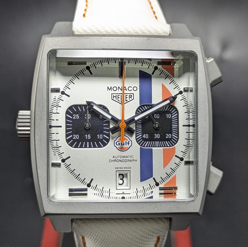 May include: A square-cased TAG Heuer Monaco chronograph watch with a silver dial, featuring two black sub-dials, a date window at the six o'clock position, and vertical blue and orange racing stripes. The watch has a white strap and orange hands.