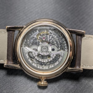Pu&ograve; includere: Un orologio da polso color oro con movimento meccanico a vista attraverso un fondello trasparente. L'orologio presenta una cassa circolare, una corona dorata testurizzata nella parte inferiore e un cinturino in pelle beige chiaro.