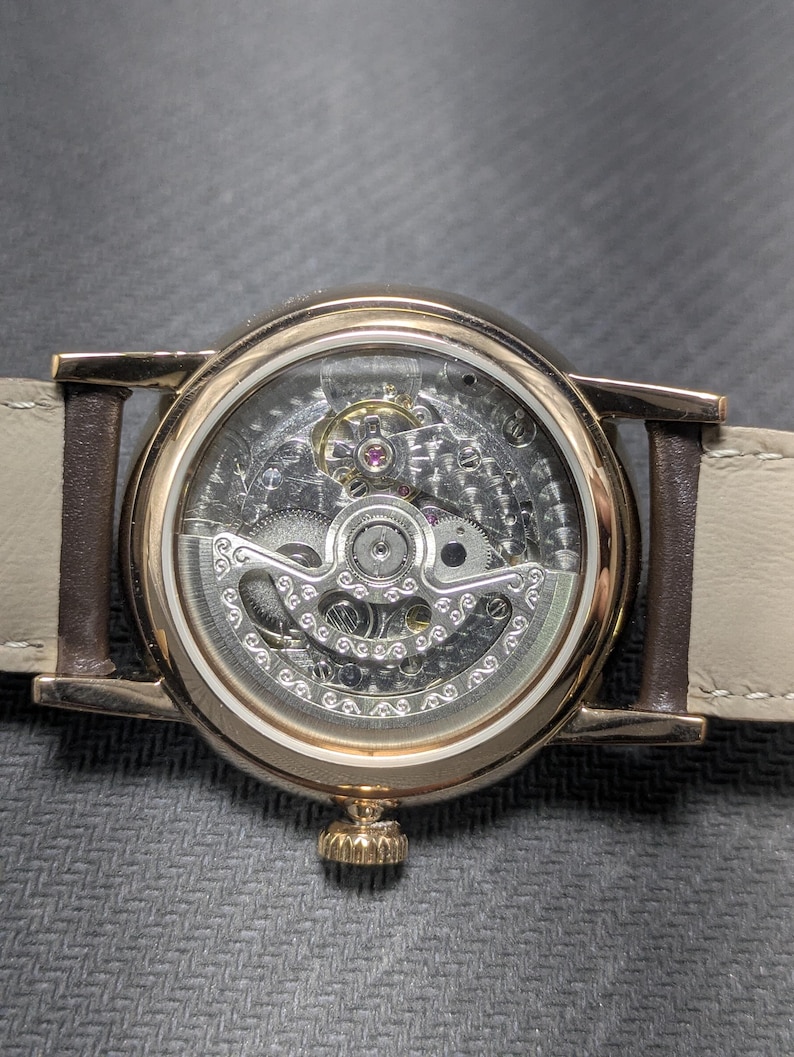 Pu&ograve; includere: Un orologio da polso color oro con movimento meccanico a vista attraverso un fondello trasparente. L'orologio presenta una cassa circolare, una corona dorata testurizzata nella parte inferiore e un cinturino in pelle beige chiaro.