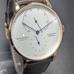 Pu&ograve; includere: Un orologio da polso Nomos Glash&uuml;tte con cassa color oro rosa, quadrante bianco e cinturino in pelle marrone. Il quadrante presenta lancette blu, un indicatore della riserva di carica Gangreserve 48 Stunden e piccoli indici neri.