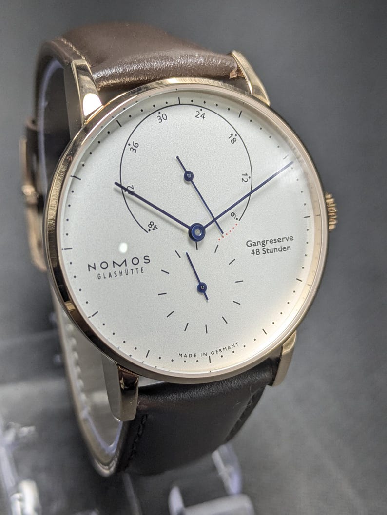 Pu&ograve; includere: Un orologio da polso Nomos Glash&uuml;tte con cassa color oro rosa, quadrante bianco e cinturino in pelle marrone. Il quadrante presenta lancette blu, un indicatore della riserva di carica Gangreserve 48 Stunden e piccoli indici neri.