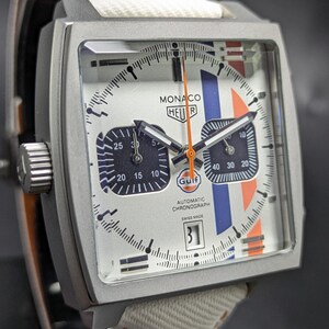 May include: A square-cased Monaco Heuer automatic chronograph watch with a white dial, blue and orange racing stripes, and a Gulf logo. The watch features two black sub-dials, a date window at the six o'clock position, and a white fabric strap.