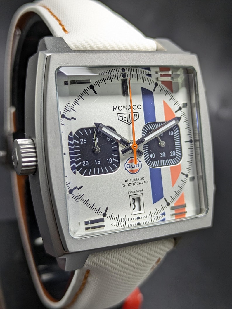 May include: A square-cased Monaco Heuer automatic chronograph watch with a white dial, blue and orange racing stripes, and a Gulf logo. The watch features two black sub-dials, a date window at the six o'clock position, and a white fabric strap.