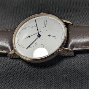 Pu&ograve; includere: Un orologio da polso rotondo con quadrante argentato, lancette sottili blu scuro e cinturino in pelle marrone. Il quadrante presenta un indicatore di riserva di carica con la scritta Gangreserve 48 Stunden, un piccolo quadrante dei secondi e Nomos Glash&uuml;tte Made in Germany.