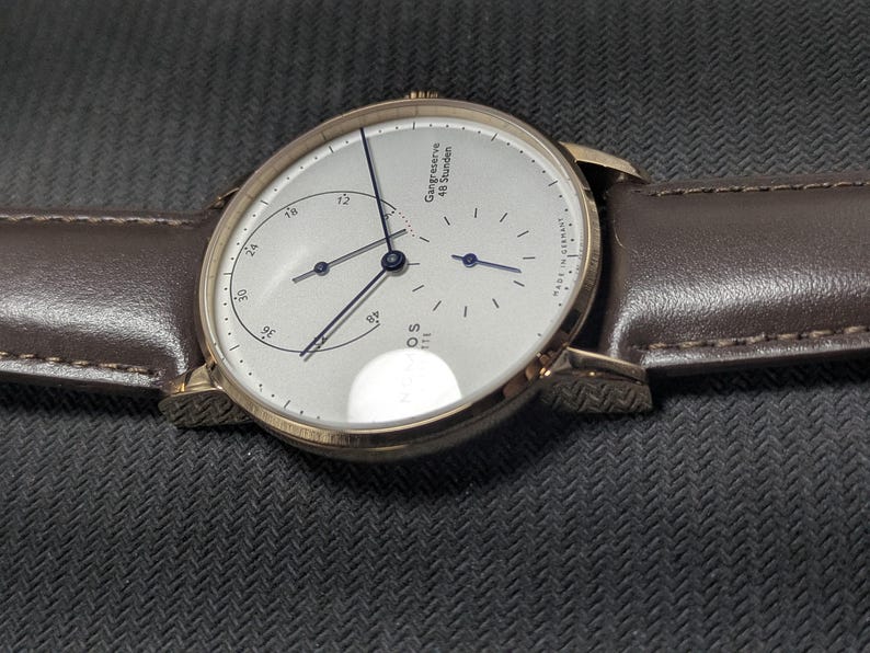 Pu&ograve; includere: Un orologio da polso rotondo con quadrante argentato, lancette sottili blu scuro e cinturino in pelle marrone. Il quadrante presenta un indicatore di riserva di carica con la scritta Gangreserve 48 Stunden, un piccolo quadrante dei secondi e Nomos Glash&uuml;tte Made in Germany.