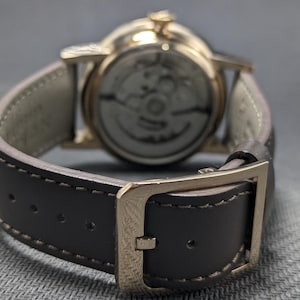 Pu&ograve; includere: Un orologio da polso con cassa in metallo color oro e cinturino in pelle marrone scuro con cuciture a vista. L'orologio presenta una chiusura a fibbia e un fondello trasparente che rivela i componenti meccanici interni del movimento.