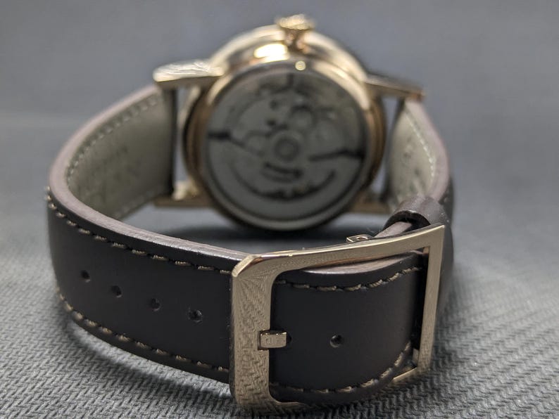 Pu&ograve; includere: Un orologio da polso con cassa in metallo color oro e cinturino in pelle marrone scuro con cuciture a vista. L'orologio presenta una chiusura a fibbia e un fondello trasparente che rivela i componenti meccanici interni del movimento.