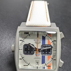 May include: A square-cased Monaco Heuer automatic chronograph watch with a silver dial, featuring blue and orange vertical racing stripes, two black sub-dials, a date window at the six o'clock position, and a white strap with orange stitching.