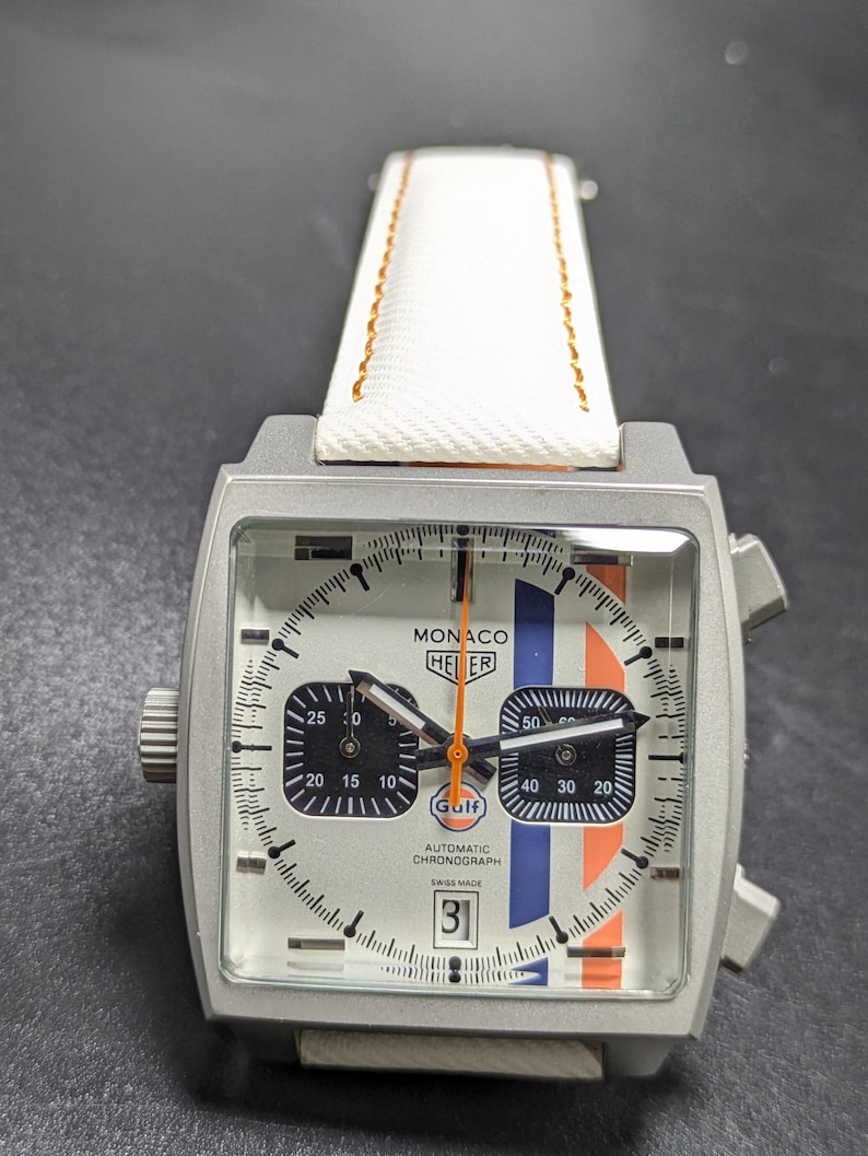 May include: A square-cased Monaco Heuer automatic chronograph watch with a silver dial, featuring blue and orange vertical racing stripes, two black sub-dials, a date window at the six o'clock position, and a white strap with orange stitching.