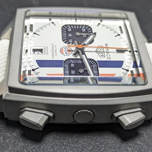 May include: A square-cased TAG Heuer Monaco chronograph watch with a white dial, blue and orange racing stripes, and a Gulf logo. The watch features a date window, two black sub-dials, a central orange hand, and a white strap with orange stitching.