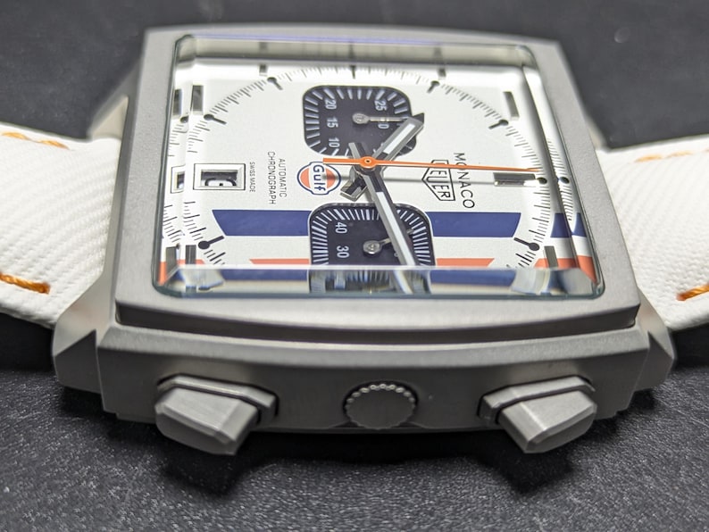 May include: A square-cased TAG Heuer Monaco chronograph watch with a white dial, blue and orange racing stripes, and a Gulf logo. The watch features a date window, two black sub-dials, a central orange hand, and a white strap with orange stitching.