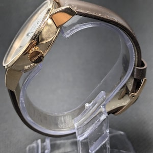 Pu&ograve; includere: Un orologio da polso con cassa in metallo color oro lucido e cinturino in pelle marrone. Il lato della cassa presenta una corona color oro testurizzata con la parola NOMOS incisa. L'orologio &egrave; esposto su un supporto in plastica trasparente.