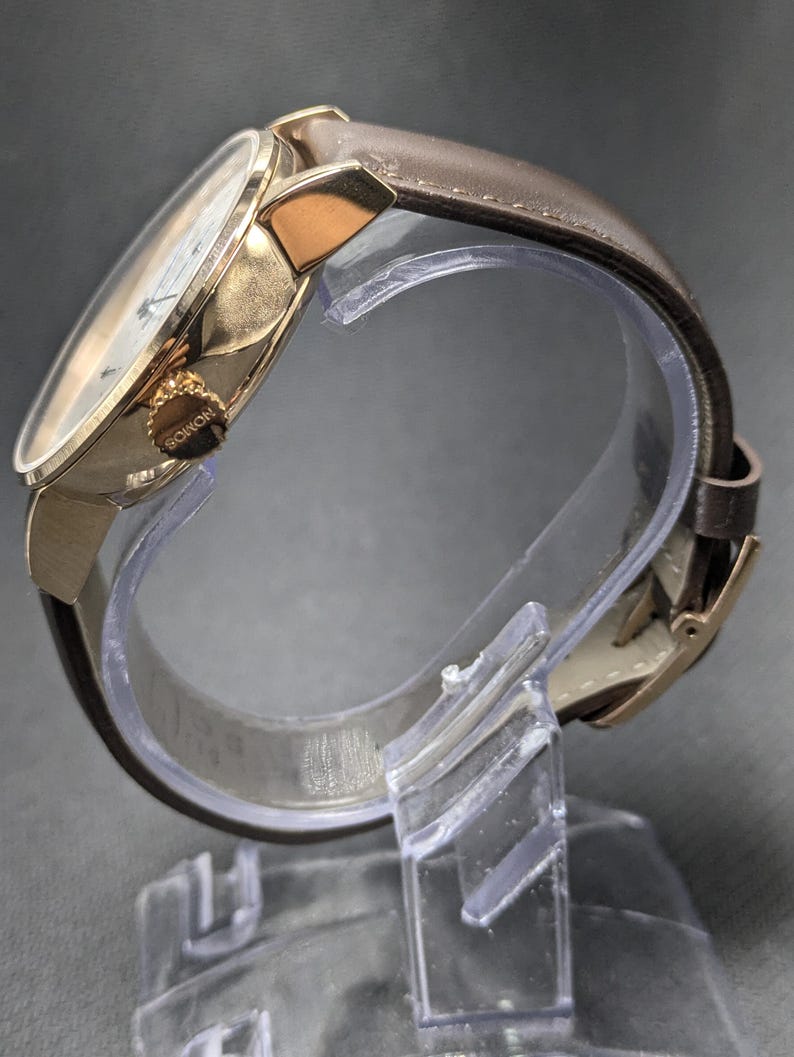 Pu&ograve; includere: Un orologio da polso con cassa in metallo color oro lucido e cinturino in pelle marrone. Il lato della cassa presenta una corona color oro testurizzata con la parola NOMOS incisa. L'orologio &egrave; esposto su un supporto in plastica trasparente.