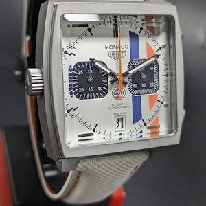May include: A square-cased TAG Heuer Monaco Gulf automatic chronograph watch with a white dial, blue and orange racing stripes, two dark sub-dials, an orange second hand, a date window at the six o'clock position, and a white fabric strap.