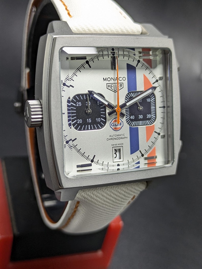 May include: A square-cased TAG Heuer Monaco Gulf automatic chronograph watch with a white dial, blue and orange racing stripes, two dark sub-dials, an orange second hand, a date window at the six o'clock position, and a white fabric strap.