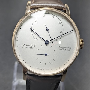 Pu&ograve; includere: Un orologio da polso Nomos Glash&uuml;tte con quadrante argentato, lancette nere sottili e cinturino in pelle marrone scuro. Il quadrante presenta un indicatore di riserva di carica con il testo Gangreserve 48 Stunden e Made in Germany in basso.