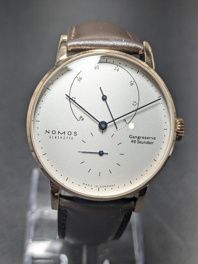 Pu&ograve; includere: Un orologio da polso Nomos Glash&uuml;tte con quadrante argentato, lancette nere sottili e cinturino in pelle marrone scuro. Il quadrante presenta un indicatore di riserva di carica con il testo Gangreserve 48 Stunden e Made in Germany in basso.