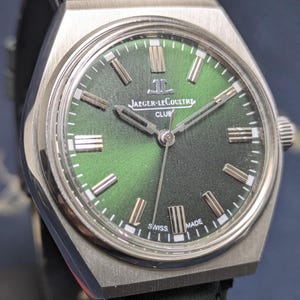 以下が含まれることがあります： グリーンの文字盤のシルバーカラーのJaeger-LeCoultre Clubウォッチのクローズアップ。時計にはシルバーの針とマーカー、グリーンのレザーストラップが付いています。時計の文字盤には「JAEGER-LECOULTRE CLUB」と「SWISS MADE」と書かれています。