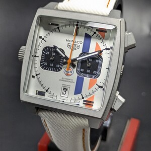 May include: A square-cased TAG Heuer Monaco automatic chronograph watch with a silver dial, two black sub-dials, and vertical blue and orange stripes. It features a white fabric strap with orange stitching, a date window at six, and Gulf branding.