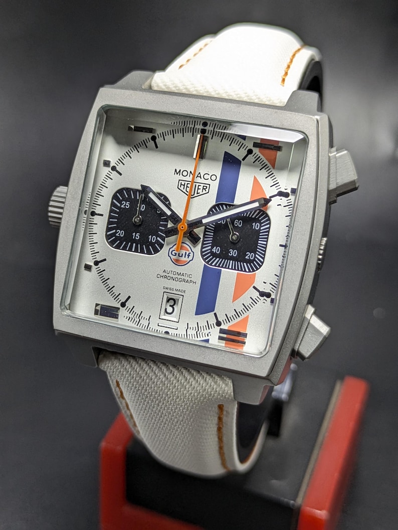 May include: A square-cased TAG Heuer Monaco automatic chronograph watch with a silver dial, two black sub-dials, and vertical blue and orange stripes. It features a white fabric strap with orange stitching, a date window at six, and Gulf branding.