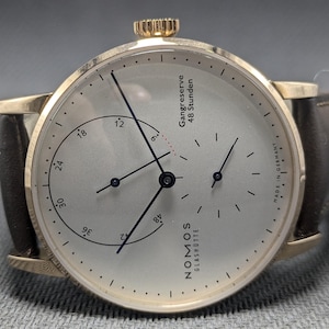 Pu&ograve; includere: Un orologio da polso color oro con quadrante argentato, cinturino in pelle marrone scuro e corona sulla parte superiore. Il quadrante presenta lancette nere, un sotto-quadrante e la scritta "NOMOS GLASH&Uuml;TTE", "Gangreserve 48 Stunden" e "MADE IN GERMANY".