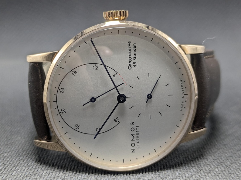 Pu&ograve; includere: Un orologio da polso color oro con quadrante argentato, cinturino in pelle marrone scuro e corona sulla parte superiore. Il quadrante presenta lancette nere, un sotto-quadrante e la scritta "NOMOS GLASH&Uuml;TTE", "Gangreserve 48 Stunden" e "MADE IN GERMANY".
