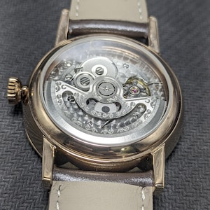 Pu&ograve; includere: Un orologio da polso meccanico color oro rosa con quadrante scheletrato che rivela intricati ingranaggi e componenti del movimento color argento. L'orologio presenta una cassa rotonda lucida, una corona testurizzata e un cinturino in pelle marrone chiaro.