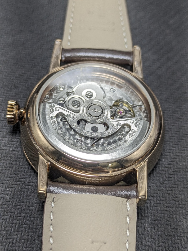 Pu&ograve; includere: Un orologio da polso meccanico color oro rosa con quadrante scheletrato che rivela intricati ingranaggi e componenti del movimento color argento. L'orologio presenta una cassa rotonda lucida, una corona testurizzata e un cinturino in pelle marrone chiaro.