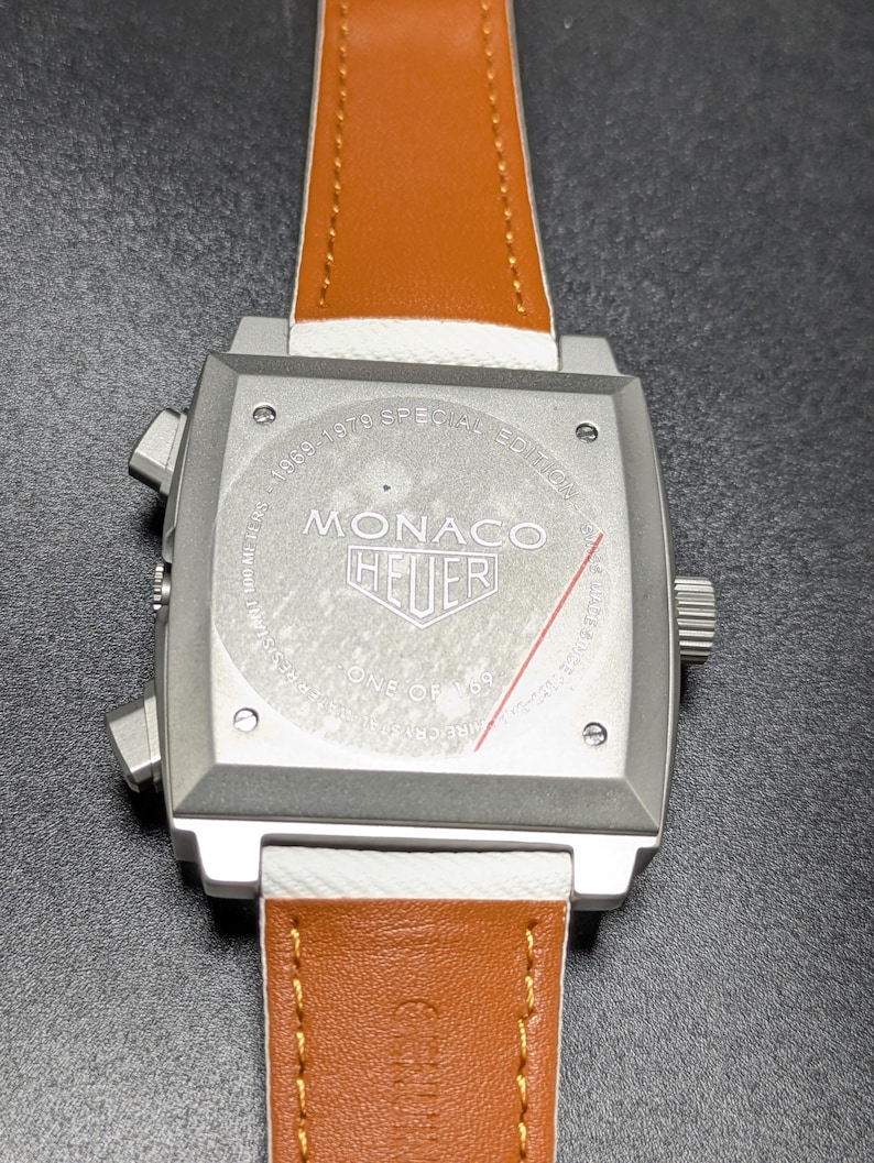 May include: A square silver-tone watch case with a tan leather strap. The case back features the text 1969-1979 SPECIAL EDITION, MONACO, HEUER, and ONE OF 169. Two pushers are on the left side, and a crown is on the right side of the watch case.