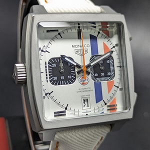 May include: A square-cased Monaco Heuer automatic chronograph watch with a white dial, two black sub-dials, and blue and orange racing stripes. The dial features the text Monaco, Heuer, Gulf, Automatic Chronograph, and Swiss Made. It has a white strap.