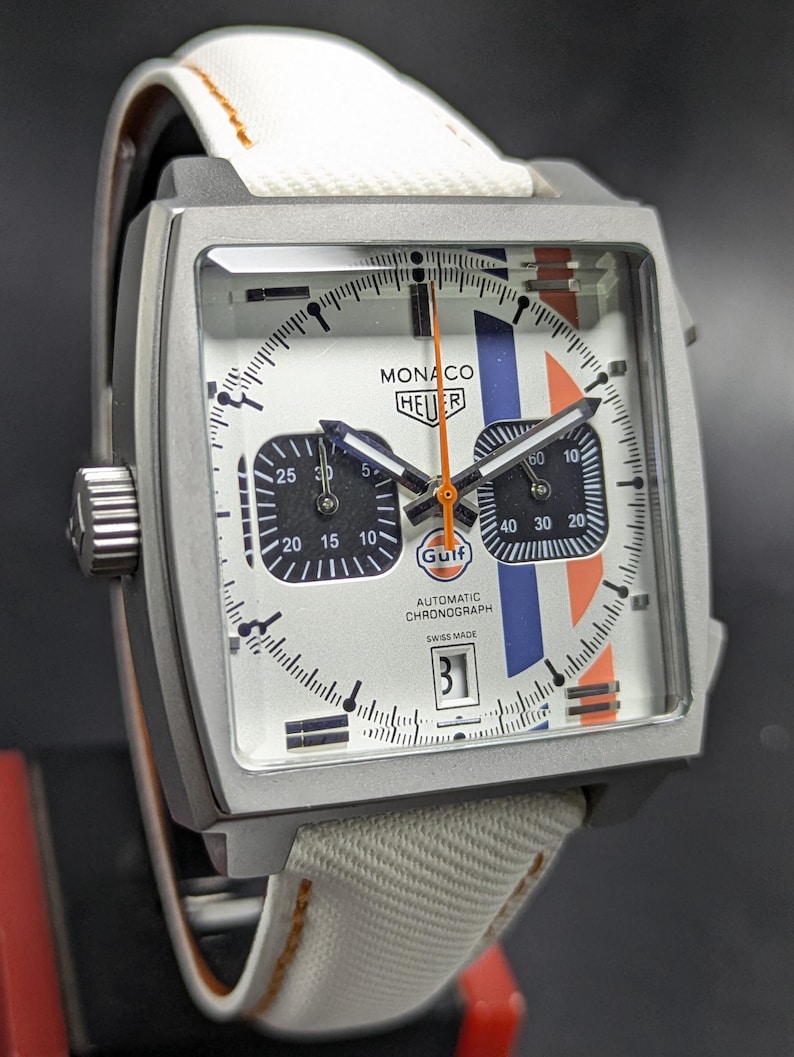 May include: A square-cased Monaco Heuer automatic chronograph watch with a white dial, two black sub-dials, and blue and orange racing stripes. The dial features the text Monaco, Heuer, Gulf, Automatic Chronograph, and Swiss Made. It has a white strap.