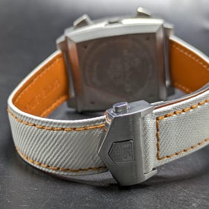 May include: A wristwatch featuring a silver-toned square case and a white textured fabric strap with orange contrast stitching. The strap includes a silver metal deployment clasp with an engraved logo, and the inner lining is tan leather.