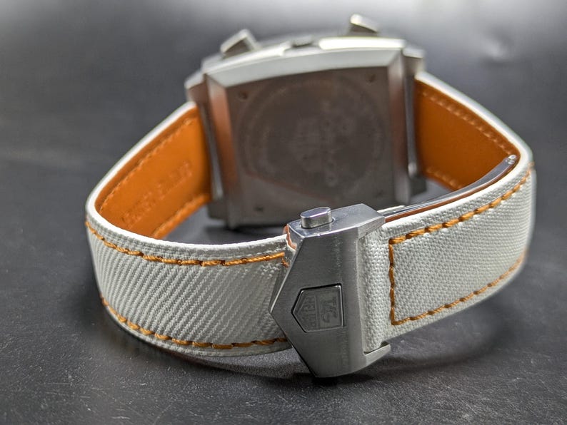 May include: A wristwatch featuring a silver-toned square case and a white textured fabric strap with orange contrast stitching. The strap includes a silver metal deployment clasp with an engraved logo, and the inner lining is tan leather.