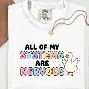 Könnte beinhalten: Weißes T-Shirt mit dem Schriftzug "ALL OF MY SYSTEMS ARE NERVOUS" in bunten Buchstaben. Neben dem Text befindet sich eine Cartoon-Gans mit ausgebreiteten Flügeln. Ein goldener Sternanhänger hängt an einer goldenen Kette.