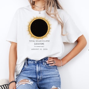 May include: A white t-shirt featuring a graphic of a total solar eclipse with a glowing yellow corona. Below the eclipse, the text reads: TOTAL SOLAR ECLIPSE, [LOCATION], THE JOHNSON FAMILY, AUGUST 12, 2026. The shirt has short sleeves and a crew neck.