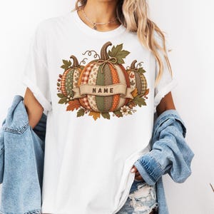 Puede incluir: Una camiseta blanca con un gráfico de tres calabazas con varios patrones de tela en colores otoñales, incluyendo cuadros, rayas y lunares. Una cinta beige sobre la calabaza central contiene el texto NAME en letras mayúsculas de color marrón oscuro.