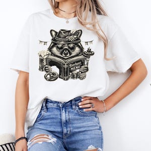 May include: A white short-sleeve t-shirt featuring a black and white graphic of a raccoon wearing a hat and sunglasses, holding a book titled Bierhaus MENU, accompanied by illustrations of a beer mug, a pretzel, and a sausage on a fork.