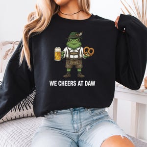 May include: A black long-sleeve sweatshirt featuring a graphic of a green frog wearing a traditional hat and lederhosen, holding a beer mug and a pretzel. Below the graphic, the white text reads WE CHEERS AT DAW in a bold, sans-serif font.