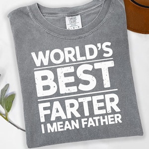 Könnte beinhalten: Graues Comfort Colors T-Shirt mit dem weißen Text "WORLD'S BEST FARTER I MEAN FATHER". Das Shirt hat einen Rundhalsausschnitt und ein Etikett mit der Aufschrift "COMFORT COLORS". Ein brauner Hut ist im Hintergrund zu sehen.