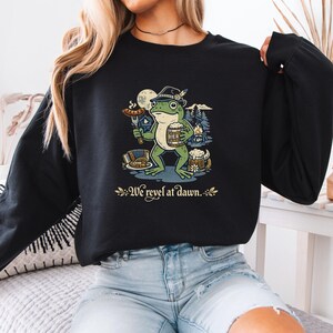 May include: A black long-sleeve sweatshirt featuring a graphic of a green frog wearing a hat, holding a sausage on a fork and a beer mug labelled BIER. The design includes a campfire, an accordion, and the text We revel at dawn. in gold.