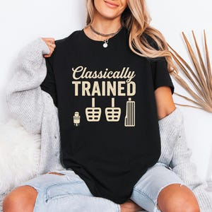 Może przedstawiać: Czarny t-shirt z napisem "Classically TRAINED" w beżowym kolorze. Poniżej beżowe ilustracje pedałów samochodowych. Koszulka ma okrągły dekolt.