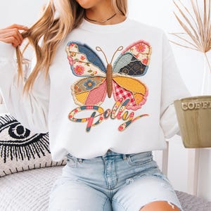 May include: A white long-sleeve sweatshirt featuring a large, colorful butterfly graphic on the chest. The butterfly wings are composed of various patchwork-style fabric patterns in shades of blue, yellow, pink, and red. Below the butterfly, the word Polly is written in a cursive, multi-colored script font that matches the patchwork aesthetic of the butterfly wings.