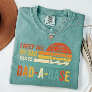 Puede incluir: Una camiseta Comfort Colors de color verde azulado con el texto "I KEEP ALL MY DAD JOKES IN A DAD-A-BASE" en naranja, amarillo y verde azulado. El gráfico presenta un diseño de disquete de estilo retro. Un collar plateado con un colgante está en la camisa.