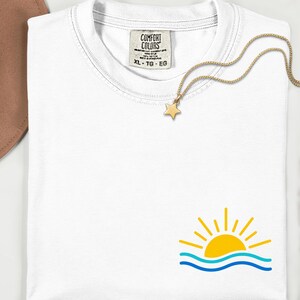 Puede incluir: Camiseta blanca con un diseño de sol amarillo sobre olas azules. Un collar con colgante de estrella dorada descansa sobre la camiseta. La etiqueta dice "COMFORT COLORS" y "XL - TG - EG".