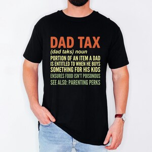 Puede incluir: Camiseta negra con el texto "DAD TAX" en naranja y blanco. El texto define "DAD TAX" como una parte de un artículo al que un padre tiene derecho cuando compra algo para sus hijos. La camiseta también menciona "PARENTING PERKS".